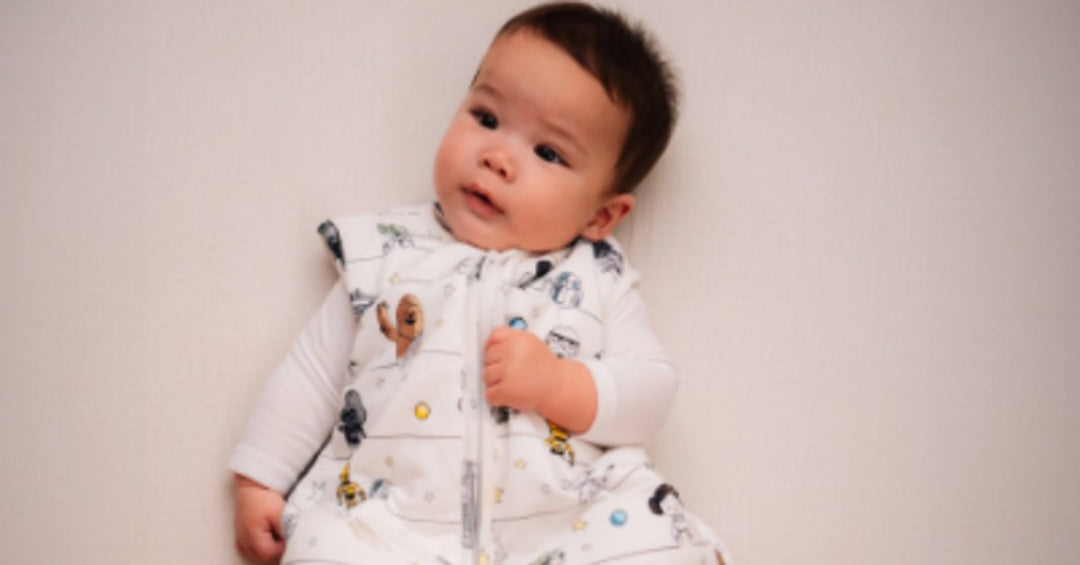 A baby lying on a white blanket while wearing a light patterned sleep sack with one arm extended outward.