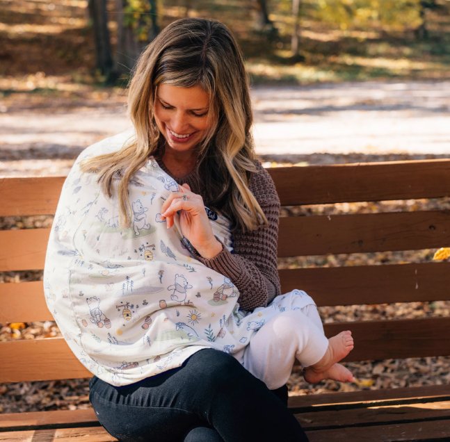 Smiling person sits on a wooden park bench holding a baby wrapped in a patterned cover, surrounded by autumn trees.