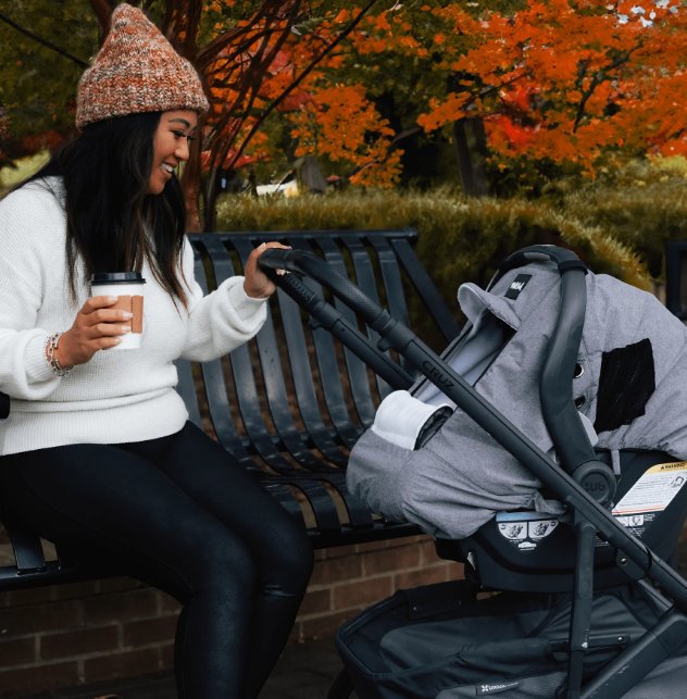 Woman in a knit hat smiles beside a stroller with a covered infant car seat while holding coffee in an autumn park.