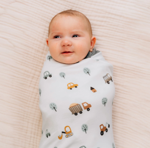 Baby swaddled in a white blanket with truck and tractor prints, lying calmly on a light quilted blanket.