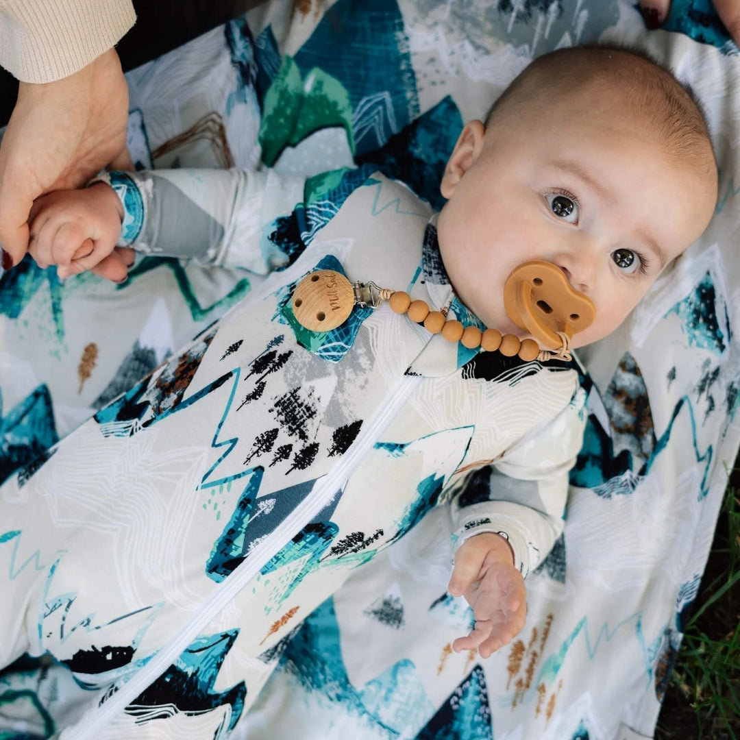 Baby wearing a blue mountain-patterned sleeper lies on matching fabric with a pacifier clipped to the outfit.