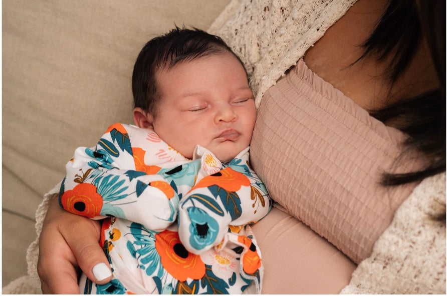 Close-up of a sleeping baby in a bright flower-patterned onesie cradled against an adult in soft neutral clothing.